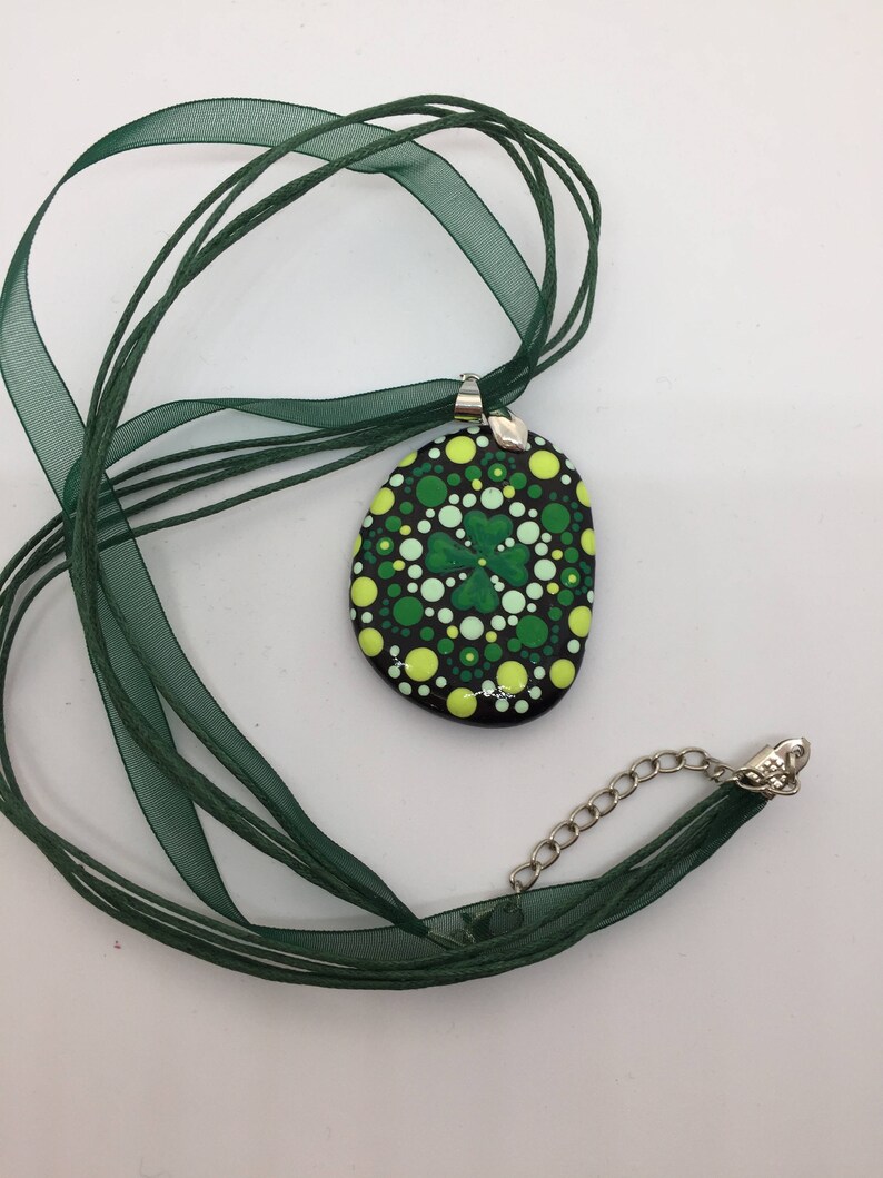 May include: A green and yellow painted rock pendant with a four-leaf clover design, hanging from a green ribbon necklace.