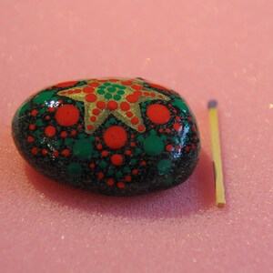 May include: A black painted rock with a gold star and red and green dots. The rock is about the size of a matchstick.