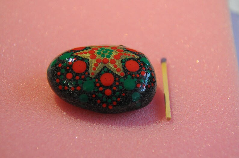 May include: A black painted rock with a gold star and red and green dots. The rock is about the size of a matchstick.