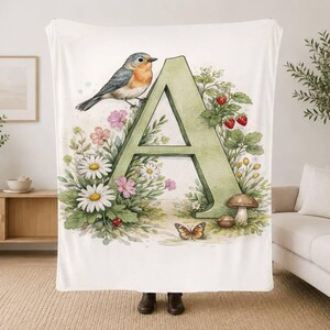 May include: A white fleece blanket featuring a large, sage green letter A decorated with a watercolour illustration of a robin, white daisies, pink flowers, red strawberries, a brown mushroom, and a small butterfly with orange and black wings.