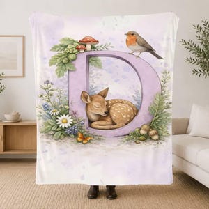 May include: A soft blanket featuring a large, light purple letter D. Inside the letter, a fawn is curled up sleeping. The design includes a robin perched on top, red mushrooms, green leaves, white daisies, blue berries, and an orange butterfly.