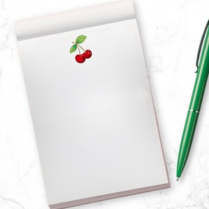 May include: A white notepad with a small red cherry illustration on the front. A green pen is lying on a white marble surface next to the notepad.