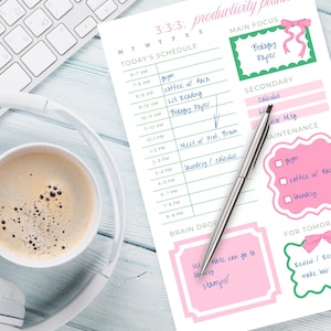 May include: A productivity planner with a pink and green design, a cup of coffee, headphones, and a keyboard. The planner includes a schedule, main focus, secondary tasks, and maintenance sections. A silver pen rests on the planner.
