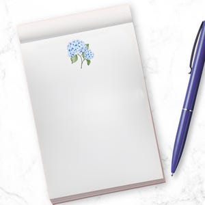 May include: A white notepad with a blue hydrangea illustration and a purple pen on a white marble surface.
