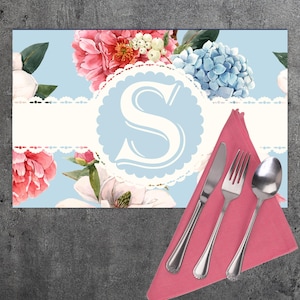 May include: A blue and white floral placemat with a white lace border and a white letter "S" in a blue circle. A pink napkin with a knife, fork, and spoon is on top of the placemat.