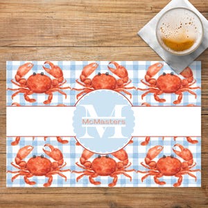 May include: A personalized placemat featuring a blue and white gingham pattern with watercolor crabs. The placemat has a light blue circle with the name "McMasters" in white. A glass of beer sits on a white napkin.