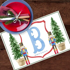 May include: A festive placemat with a watercolor design featuring a blue initial "B" and the name "Bronson" in a red crest. Two toy soldier nutcrackers and Christmas trees flank the initial. A red plate and blue plate with silverware and a napkin are in the upper left corner.