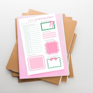 May include: A pink productivity planner with the text "3:3:3: productivity planner" and a daily schedule layout. The planner includes sections for "Main Focus," "Secondary," "Maintenance," "Brain Drop," and "For Tomorrow." The planner is stacked on top of several brown notebooks.