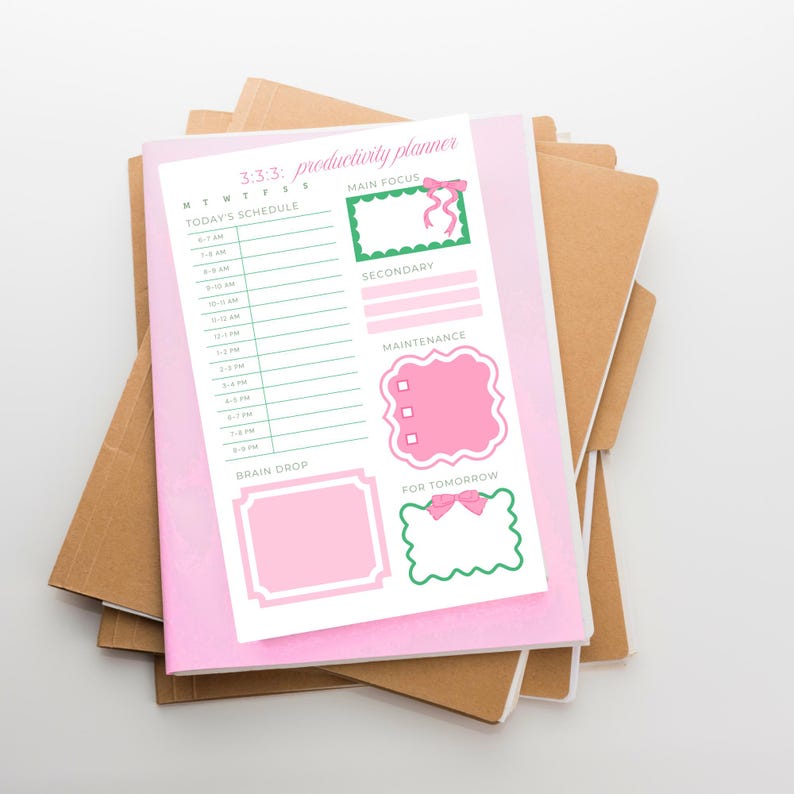 May include: A pink productivity planner with the text "3:3:3: productivity planner" and a daily schedule layout. The planner includes sections for "Main Focus," "Secondary," "Maintenance," "Brain Drop," and "For Tomorrow." The planner is stacked on top of several brown notebooks.