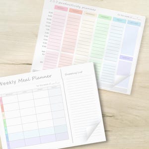 May include: Two white paper planners with pastel color-coded sections. The top planner is labeled "3:3:3 productivity planner" and the bottom planner is labeled "Weekly Meal Planner" with a "Shopping List" section. Both have sections for the week.