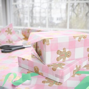 Peut inclure: Deux cadeaux emball&eacute;s sur une table recouverte de papier cadeau &agrave; carreaux roses et blancs, orn&eacute; de bonhommes en pain d'&eacute;pices. Des ciseaux et un ruban vert sont &eacute;galement visibles, pr&egrave;s d'une fen&ecirc;tre.
