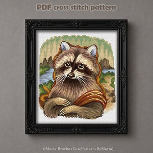 May include: Cross-stitch pattern featuring a raccoon in a framed portrait. The raccoon has brown and gray fur, with a black mask and a Mona Lisa-like pose. The background shows a landscape with a river and trees. The frame is black and ornate. Text at the top reads "PDF cross stitch pattern".