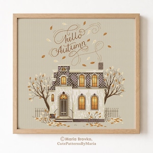 May include: A cross-stitch pattern of a house with the text "Hello Autumn" and falling leaves. The house is white with a dark roof and lit windows, framed in a wooden square.