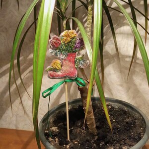 May include: A cross-stitch design of a red rubber boot filled with colorful flowers, attached to a wooden stick and placed in a potted plant.