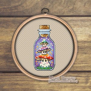 May include: Cross-stitch design of three colorful mushrooms inside a glass bottle with a cork stopper. The bottle is set against a light beige background within a wooden embroidery hoop. The text "MARIA BROVKO" is at the bottom.