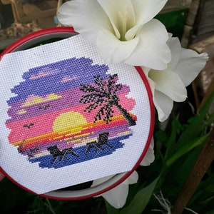 Puede incluir: Un bordado de punto de cruz en un bastidor circular rojo que muestra una puesta de sol con un sol amarillo, nubes rosas y moradas, una palmera negra y dos tumbonas negras en una playa azul sobre un fondo de tela blanca.