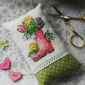 May include: A white fabric pillow with a cross-stitch design of a red rain boot filled with colorful flowers. The pillow has a green and white polka dot fabric backing and a white lace trim.