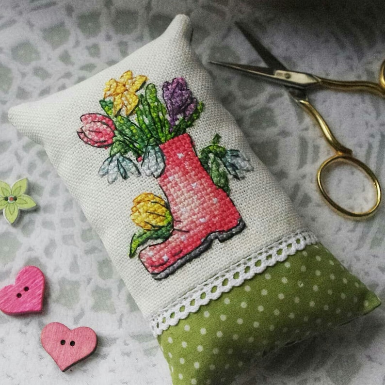 May include: A white fabric pillow with a cross-stitch design of a red rain boot filled with colorful flowers. The pillow has a green and white polka dot fabric backing and a white lace trim.