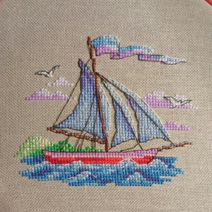 May include: A cross-stitch embroidery of a red sailboat with blue sails and a pink and white flag. The sailboat is sailing on blue water with white waves. There are pink and white clouds in the background.