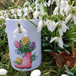 May include: A white canvas bag with a cross-stitch design of a pink rain boot filled with colorful flowers. The bag is sitting on a bed of green grass and white snowdrop flowers.