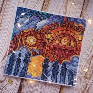 May include: A cross-stitch embroidery depicting a whimsical, colorful building with a large, round window and a red door. The building is surrounded by a swirling, blue and purple sky with clouds. There are six figures standing in front of the building, all wearing dark clothing.