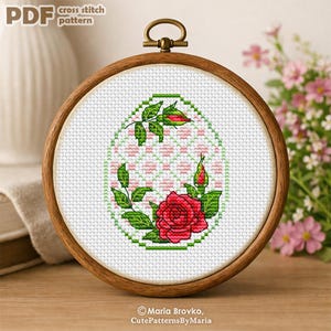 Puede incluir: Un patrón de punto de cruz en un aro de madera presenta una rosa roja con hojas y capullos verdes. El diseño está sobre un fondo blanco con un patrón geométrico rosa y verde. El texto "PDF cross stitch pattern" está en la parte superior.