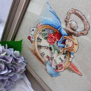 以下が含まれることがあります： アリス・イン・ワンダーランドのクロスステッチ刺繍。アリスは白いウサギ、フラミンゴ、ティーカップと一緒に懐中時計の上で眠っています。時計の文字盤はローマ数字で、背景は薄いベージュのリネンです。
