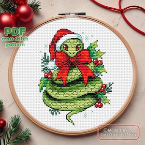 May include: A cross-stitch pattern of a green snake wearing a Santa hat and a red bow. The snake is surrounded by holly and berries, set in a wooden embroidery hoop. The text "PDF cross stitch pattern" is in the upper left.