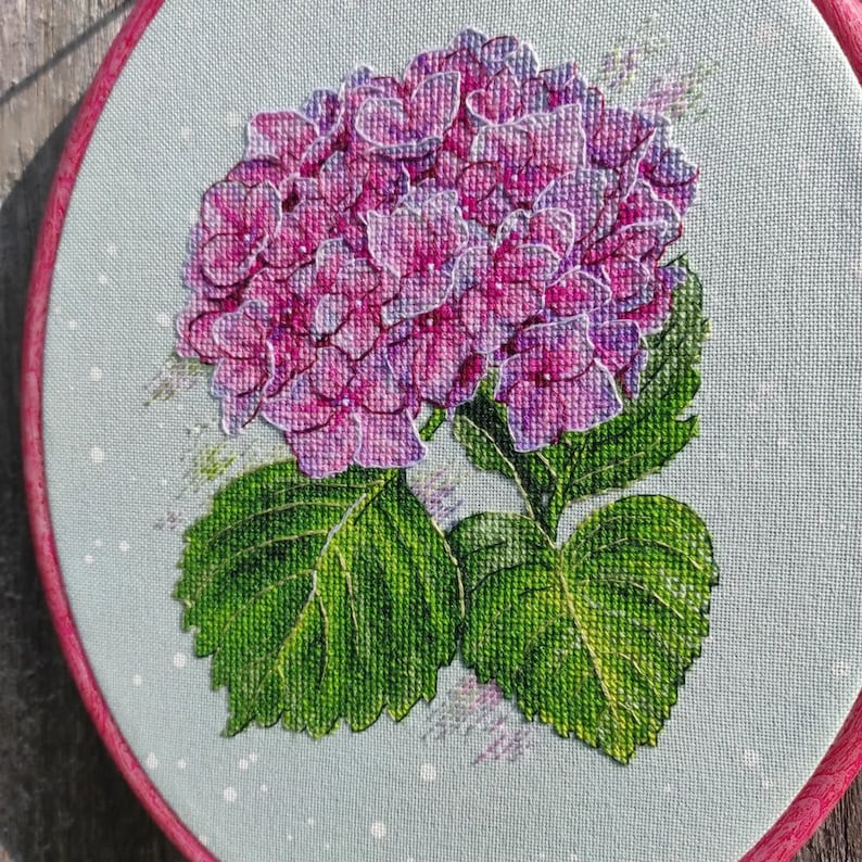 May include: A close-up of a pink and purple hydrangea flower with green leaves, embroidered on a light blue fabric.