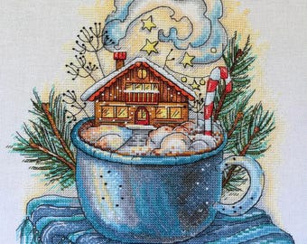 Puede incluir: Una taza azul con una pequeña casa y un bastón de caramelo dentro, rodeada de nieve y una rama de pino. La taza está sentada sobre una bufanda a rayas azul y blanca. La casa tiene un techo rojo y ventanas con luces amarillas.