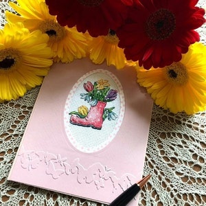May include: A pink card with a cross-stitch design of a red rain boot filled with colorful flowers. The card has a decorative border of embossed flowers. A pen is lying on the card.