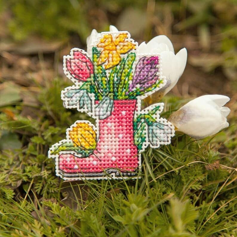 May include: A pink rubber boot filled with a bouquet of yellow daffodils, purple crocuses, and red tulips. The boot is sitting on a bed of green grass.