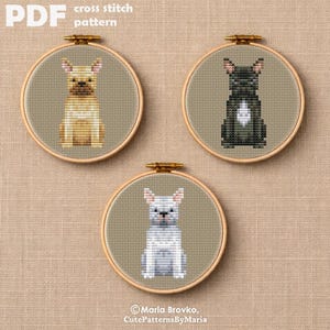Puede incluir: Tres patrones de punto de cruz de perros en aros de bordado. Los perros son en tonos beige, negro y blanco. El aro superior izquierdo tiene un perro beige, el superior derecho tiene un perro negro y el inferior tiene un perro blanco. El texto "PDF cross stitch pattern" está en la parte superior.