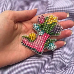 May include: A pink rubber boot filled with a bouquet of colorful flowers, including yellow tulips, purple crocuses, and white snowdrops. The boot is embroidered with a white polka dot pattern.