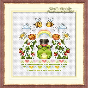 Op de afbeelding: Borduurpatroon met een groene kikker die een hoge hoed draagt, omringd door aardbeien, bloemen en bijen. Het patroon bevat een regenboog en de tekst "Maria Brovko (CutePatternsByMaria)".