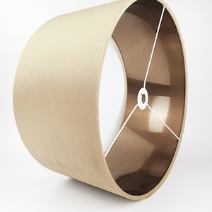 K&ouml;nnte beinhalten: Ein beigefarbener Lampenschirm mit goldfarbener Metallic-Futter. Der Lampenschirm ist zylindrisch und hat eine glatte, samtige Textur.