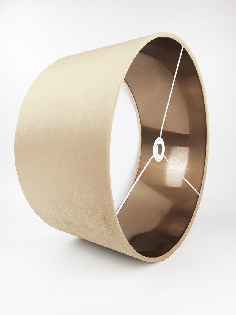 K&ouml;nnte beinhalten: Ein beigefarbener Lampenschirm mit goldfarbener Metallic-Futter. Der Lampenschirm ist zylindrisch und hat eine glatte, samtige Textur.