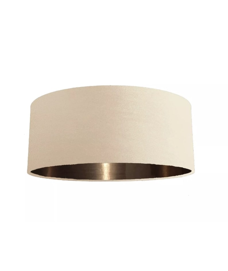 K&ouml;nnte beinhalten: Ein cremefarbener Lampenschirm mit goldfarbenem Metallic-Besatz. Der Lampenschirm hat eine zylindrische Form.