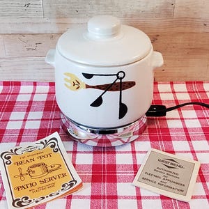 May include: A vintage white stoneware bean pot with a lid, featuring a decorative design of utensils. The pot sits on a chrome base with a power cord, placed on a red and white checkered tablecloth. Two informational cards are also present.