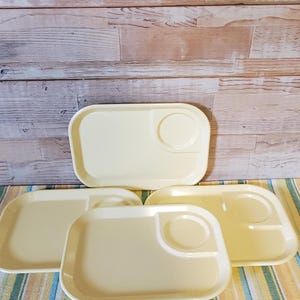 May include: A collection of five vintage, pale yellow plastic TV trays. Each tray has a rectangular shape with rounded corners and a circular indentation for a cup or glass. The trays are arranged on a striped tablecloth.