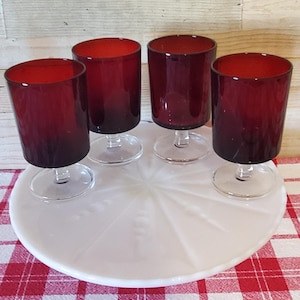 May include: Four burgundy-colored wine glasses with clear stems are arranged on a white, circular serving tray. The tray sits on a red and white checkered tablecloth, with a light wood background.