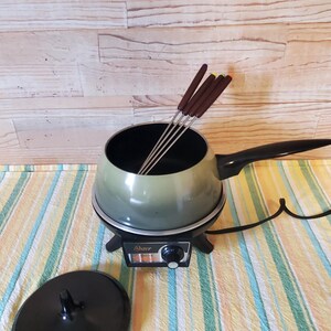 May include: An electric fondue pot with a sage green exterior and a black handle. The base features a black control panel with the Oster brand name and a dial. Four metal fondue forks with brown handles are inside the pot, and a black lid sits nearby.