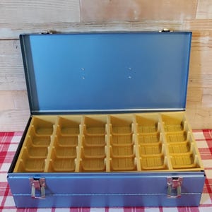 May include: A rectangular blue metal storage case with a hinged lid and two silver-toned metal latches on the front. The interior features a removable yellow plastic insert with twenty-four individual compartments arranged in a grid pattern.