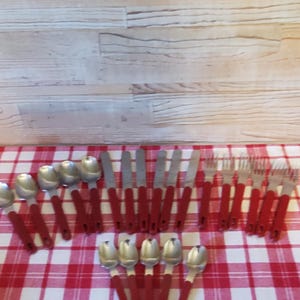 May include: A collection of stainless steel flatware featuring red plastic handles, including four spoons, four knives, four forks, and five smaller spoons, all arranged in a group on a red and white checkered surface.