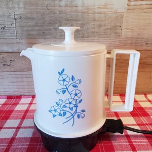 May include: White electric coffee pot with a blue floral design. The pot has a white lid and handle, and sits on a black base. The pot is on a red and white checkered tablecloth.