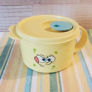 May include: Yellow plastic bowl with a lid and handle. The bowl features a cartoon face on the front and a light blue knob on the lid. The bowl is sitting on a striped fabric.
