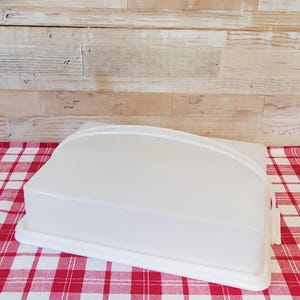 May include: A white plastic food storage container with a curved top and a rectangular base. The container is on a red and white checkered tablecloth. The background is a light brown wooden surface.