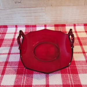 May include: A vintage, ruby red glass serving dish with two ornate handles. The dish has a unique shape and a black trim. It sits on a red and white checkered tablecloth.