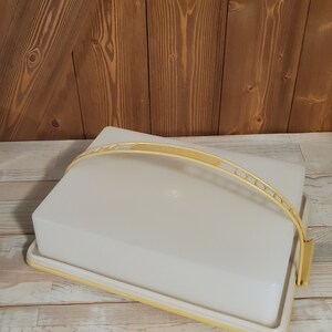 May include: A rectangular, off-white plastic cake carrier with a pale yellow handle and trim. The handle is arched and has a decorative design. The carrier sits on a light-colored wooden surface with a darker wooden background.