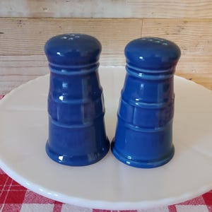 May include: A pair of matching cobalt blue ceramic salt and pepper shakers. Each shaker features a cylindrical body with a rounded top, a series of horizontal ridges around the middle, and a flared base. The tops have small holes for dispensing.